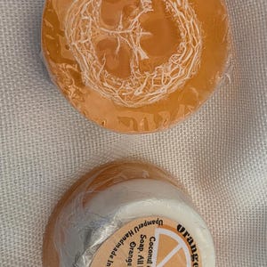 May include: Two round bars of orange loofah soap wrapped in clear plastic. The top bar shows a textured loofah embedded in translucent orange soap. The bottom bar has a white base and a circular label reading Orange Loofah, Coconut & Shea Butter Soap, All Natural Loofa, Orange Essential oil, UpamperU Handmade in Greeley Co. (970) 371-6141.