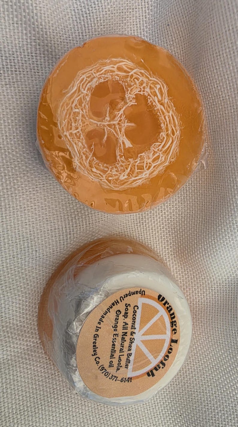 May include: Two round bars of orange loofah soap wrapped in clear plastic. The top bar shows a textured loofah embedded in translucent orange soap. The bottom bar has a white base and a circular label reading Orange Loofah, Coconut & Shea Butter Soap, All Natural Loofa, Orange Essential oil, UpamperU Handmade in Greeley Co. (970) 371-6141.