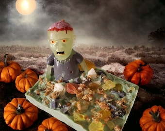 May include: A green zombie figurine is partially submerged in a green and brown candy bar with various toppings. The candy bar is surrounded by small orange pumpkins.
