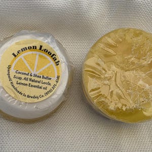 May include: Two round bars of yellow soap wrapped in clear plastic. One bar features a circular label with a lemon graphic and text reading Lemon Loofah, Coconut & Shea Butter Soap, All Natural Loofa, Lemon Essential oil, UpamperU Handmade In Greeley Co. (970) 371-6141.