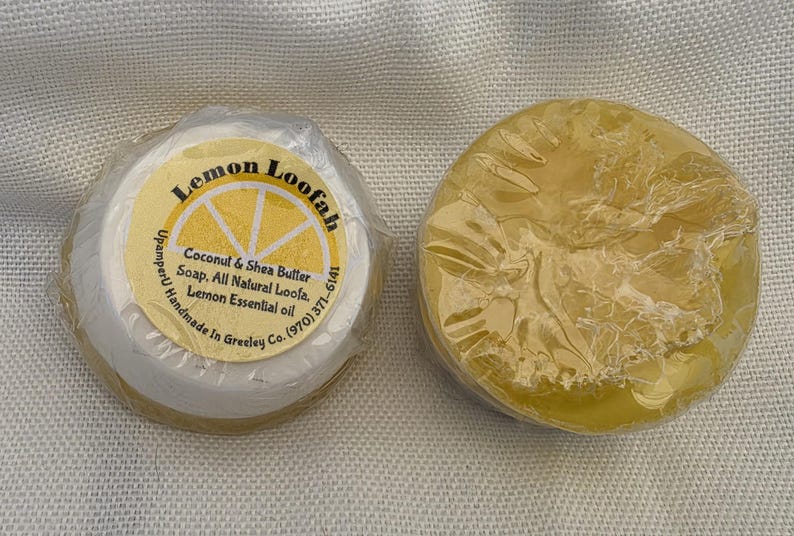 May include: Two round bars of yellow soap wrapped in clear plastic. One bar features a circular label with a lemon graphic and text reading Lemon Loofah, Coconut & Shea Butter Soap, All Natural Loofa, Lemon Essential oil, UpamperU Handmade In Greeley Co. (970) 371-6141.