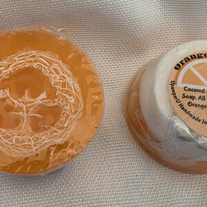 May include: Two round bars of soap wrapped in clear plastic. One bar is translucent orange with a visible loofah fiber embedded in the center. The other bar is white and orange with a circular label reading Orange Loofah, Coconut & Shea Butter Soap, All Natural Loofa, Orange Essential oil, UpamperU Handmade In Greeley Co. (970) 371-6141.