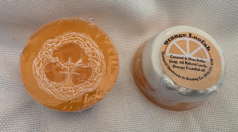 May include: Two round bars of soap wrapped in clear plastic. One bar is translucent orange with a visible loofah fiber embedded in the center. The other bar is white and orange with a circular label reading Orange Loofah, Coconut & Shea Butter Soap, All Natural Loofa, Orange Essential oil, UpamperU Handmade In Greeley Co. (970) 371-6141.