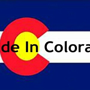 May include: A rectangular flag featuring horizontal blue and white stripes with a large red letter C containing a yellow circle in the center. The black text Made In Colorado is printed horizontally across the white stripe.