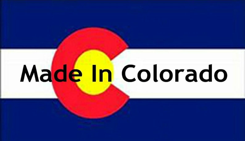 May include: A rectangular flag featuring horizontal blue and white stripes with a large red letter C containing a yellow circle in the center. The black text Made In Colorado is printed horizontally across the white stripe.