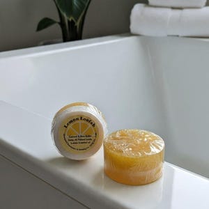 May include: Two round, yellow lemon loofah soaps wrapped in clear plastic. One soap shows a circular label with the text Lemon Loofah, Centered A Shea Butter Soap, All Natural Loods, Lemon Essential oil, and Upward Ingredients in Greeley Co.