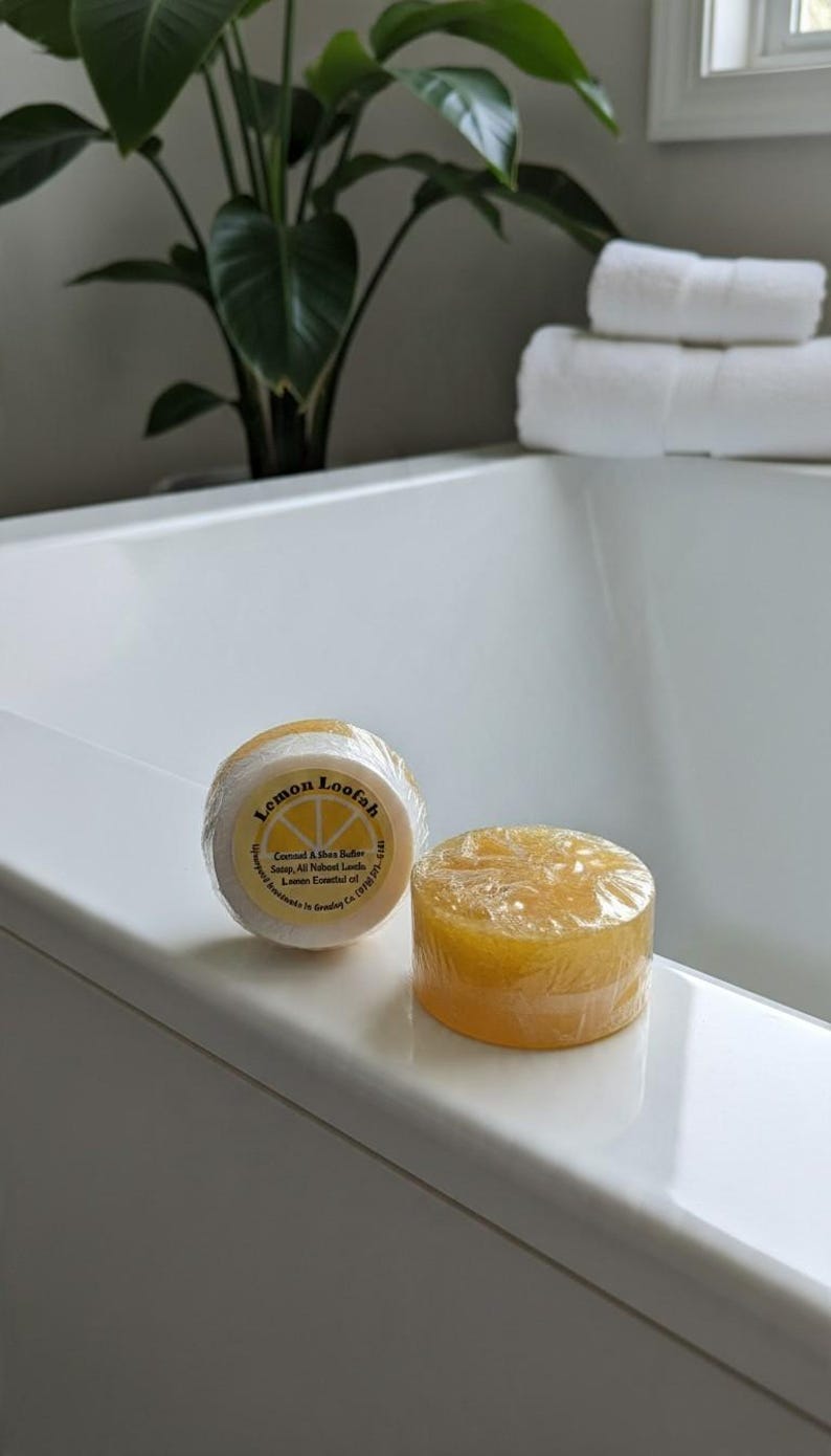 May include: Two round, yellow lemon loofah soaps wrapped in clear plastic. One soap shows a circular label with the text Lemon Loofah, Centered A Shea Butter Soap, All Natural Loods, Lemon Essential oil, and Upward Ingredients in Greeley Co.