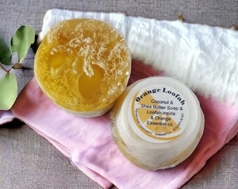 May include: A round, translucent yellow soap bar containing a fibrous loofah, paired with a smaller, opaque white soap bar featuring a circular label that reads Orange Loofah, Coconut & Shea Butter Soap & Loofah inside & Orange Essential oil.