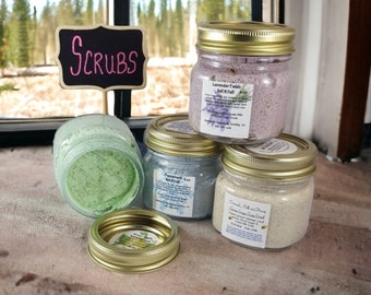 May include: Four glass jars with gold lids filled with different colored bath scrubs. The jars are labeled with the names of the scrubs: Lavender Fields Bath Salt, Peppermint & Tea Bath Salt, Coconut Milk and Honey Green Sugar Scrub. A black chalkboard sign reads "Scrubs".