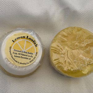 May include: Two round bars of yellow soap wrapped in clear plastic. One bar features a circular label that reads Lemon Loofah, Coconut & Shea Butter Soap, All Natural Loofa, Lemon Essential oil, UpamperU Handmade In Greeley Co. (970) 371-6141.