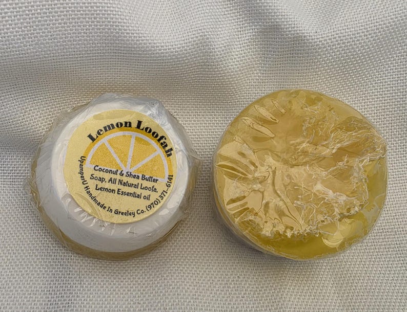 May include: Two round bars of yellow soap wrapped in clear plastic. One bar features a circular label that reads Lemon Loofah, Coconut & Shea Butter Soap, All Natural Loofa, Lemon Essential oil, UpamperU Handmade In Greeley Co. (970) 371-6141.