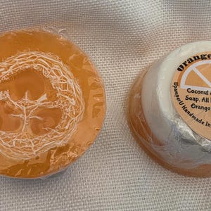 May include: Two round bars of soap wrapped in clear plastic. One bar is translucent orange with a visible loofah fiber embedded in the center. The other bar is white and orange with a circular label reading Orange Loofah, Coconut & Shea Butter Soap, All Natural Loofa, Orange Essential oil, UpamperU Handmade In Greeley Co. (970) 371-6141.
