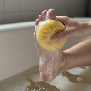 May include: A round, yellow, textured loofah sponge covered in white soap suds, being used to scrub the sole of a foot. The sponge has a fibrous, porous surface and is held firmly against the skin, creating a lather of bubbles.