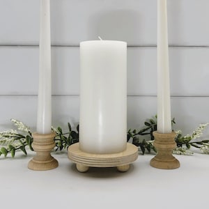 May include: A set of three white candles on wooden candle holders. The candles are arranged in a triangle shape. The candles are on a white wooden surface.