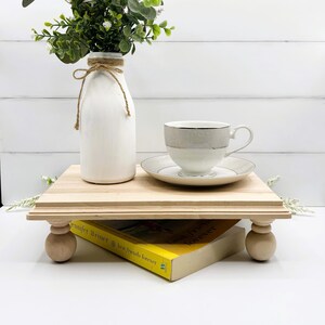 May include: A wooden tiered stand with turned legs, featuring a white milk bottle vase with a brown twine bow, filled with green and white faux greenery, a white teacup and saucer, and a yellow paperback book titled "Best Friends Forever" by Jennifer Weiner.
