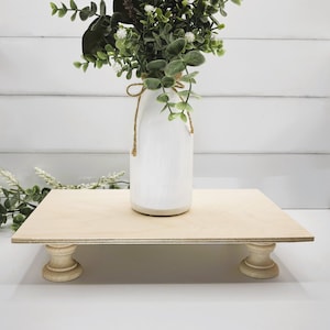 May include: A white painted milk glass vase with a brown twine bow, filled with a bouquet of green eucalyptus and white flowers, sits on a wooden pedestal with turned legs.