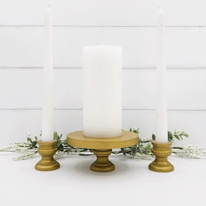 Puede incluir: Un conjunto de tres candelabros dorados con velas blancas. El candelabro central es un pedestal con una base redonda. Los dos candelabros laterales son más pequeños y tienen un diseño más tradicional.