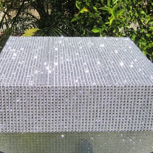 Pode incluir: Um suporte de bolo quadrado coberto com glitter prateado. O suporte é feito de papelão e tem uma superfície lisa e brilhante.
