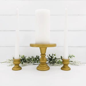 Puede incluir: Una composición decorativa con tres velas blancas. Una gran vela de pilar se asienta sobre un pedestal dorado, flanqueada por dos velas cónicas delgadas en soportes dorados a juego. La vegetación añade un toque natural.