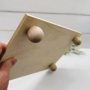 Puede incluir: Una bandeja de madera con cuatro patas redondas de madera. La bandeja es de color marrón claro y tiene una superficie lisa.