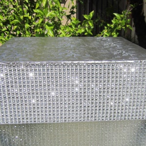 Pode incluir: Uma caixa retangular prateada com uma parte superior estampada e uma borda brilhante coberta de strass. A caixa está sobre uma superfície de vidro, com folhagem verde ao fundo. A caixa é provavelmente usada para armazenamento ou exibição.