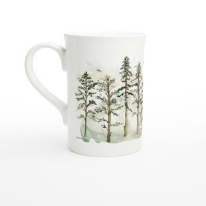 May include: White ceramic mug featuring a watercolour design of a line of green trees in a misty forest.