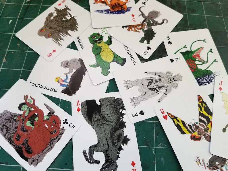 May include: A deck of playing cards featuring various monster illustrations. The cards are scattered across a green surface, showcasing the artwork on each card. The designs include a variety of creatures, with suits and card values visible.