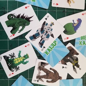 May include: A collection of playing cards featuring illustrations of various monsters and creatures. The cards have a white background with colorful images and suit symbols. Some cards display the text "DIE KAIJU!" and "KAIJU!".