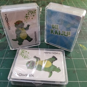 May include: Three decks of playing cards in clear plastic cases. Two decks feature a green cartoon dinosaur labeled "JOKER". The third deck has "DIE KAIJU!" on a blue background. The cards are on a green surface.