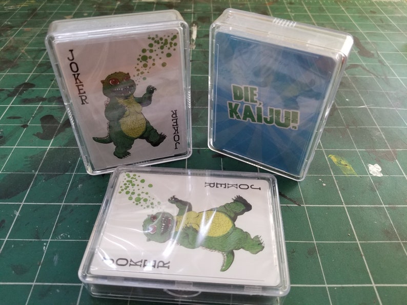 May include: Three decks of playing cards in clear plastic cases. Two decks feature a green cartoon dinosaur labeled "JOKER". The third deck has "DIE KAIJU!" on a blue background. The cards are on a green surface.