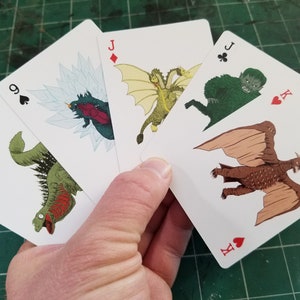 May include: A hand holding a fan of playing cards featuring monster illustrations. The cards show various monsters, including a three-headed dragon and a winged creature. The cards are white with black suit symbols and red face values.