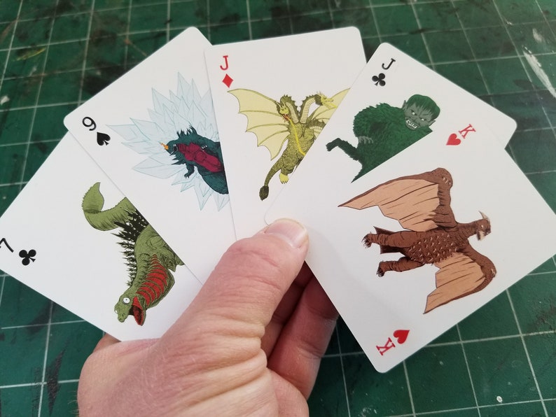 May include: A hand holding a fan of playing cards featuring monster illustrations. The cards show various monsters, including a three-headed dragon and a winged creature. The cards are white with black suit symbols and red face values.