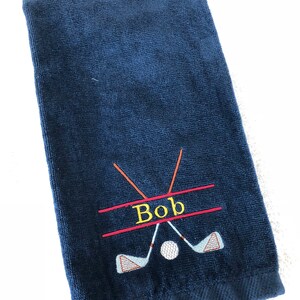 May include: A navy blue golf towel with a personalized name "Bob" embroidered in yellow thread. Two crossed golf clubs are embroidered in red and orange thread with a white golf ball in the center.