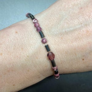 May include: A bracelet with pink and white patterned beads and a dark purple crystal bead. The bracelet is made with dark grey metal beads.
