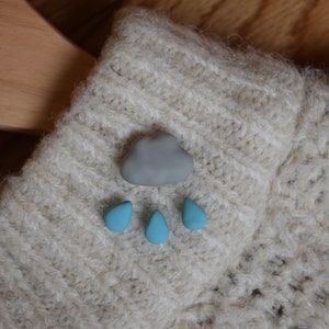 May include: A gray cloud with three blue raindrops on a beige knitted fabric.