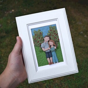 May include: A white frame with a 3D clay sculpture of a couple standing in front of two trees. The couple is holding hands and the trees have red berries.
