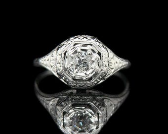 Op de afbeelding: Een witte metalen ring met een centrale diamant, gezet in een achthoekige setting. De ring heeft gedetailleerde filigraanpatronen. De afbeelding toont de ring tegen een zwarte achtergrond, met zijn weerspiegeling eronder.
