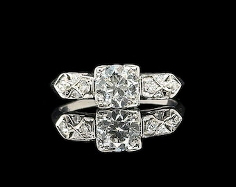 Könnte beinhalten: Ein Platinring mit einem großen, quadratisch geschliffenen Diamanten als Mittelstein. Der Ring hat kleinere Diamanten auf den Schultern und eine reflektierende Oberfläche darunter. Der Ring ist vor einem schwarzen Hintergrund platziert.