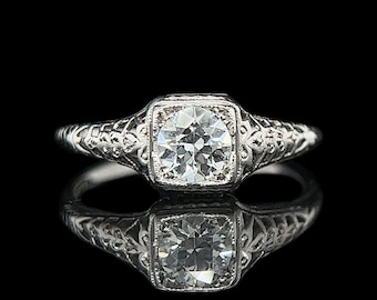 May include: A vintage engagement ring with a single round diamond set in a square bezel. The band is decorated with a delicate floral design.
