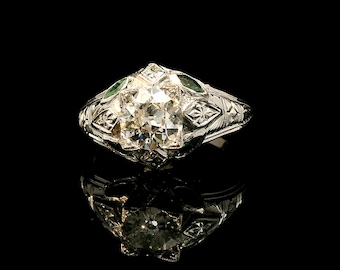 May include: A vintage-style ring with a large, square-cut diamond as the centerpiece. The ring is crafted from a silver-colored metal and features intricate filigree and small green accents. The ring is set against a black background.