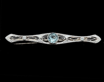 May include: An antique bar brooch featuring a central aquamarine gemstone and two smaller accent stones. The filigree design is detailed, set against a black backdrop. The metal is likely silver or white gold, showcasing a classic aesthetic.