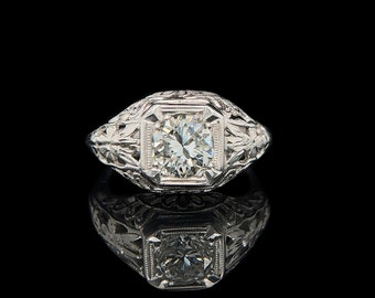 Könnte beinhalten: Ein detaillierter Weißgoldring mit einem großen, runden, klaren Diamanten, der in einem quadratischen Rahmen gefasst ist. Der Ring weist auf dem Band und den Schultern aufwendige Blumen- und Blattgravuren auf. Der Ring wird auf einer reflektierenden schwarzen Oberfläche präsentiert.