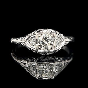 Puede incluir: Un anillo de compromiso vintage con un diamante redondo brillante central rodeado de diamantes más pequeños. El anillo está engastado en platino y presenta un diseño filigranado intrincado.