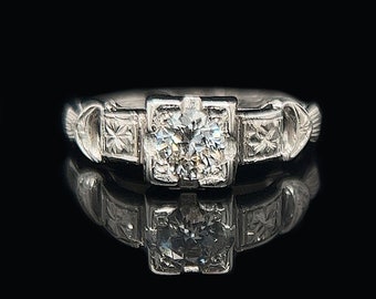May include: A silver ring with a large, round, clear diamond center stone. The ring has a square setting with smaller accent stones on each side. The band features decorative detailing and a reflective surface.