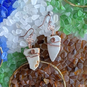 May include: A pair of earrings made with white ceramic pieces wrapped with copper wire. The earrings are displayed on a bed of brown, white, and green sea glass.