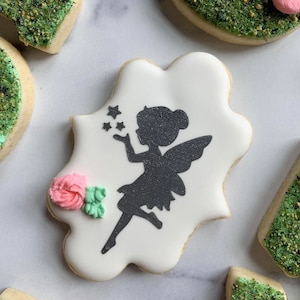 May include: A white sugar cookie decorated with a black silhouette of a fairy blowing stars. The cookie is decorated with pink and green frosting.