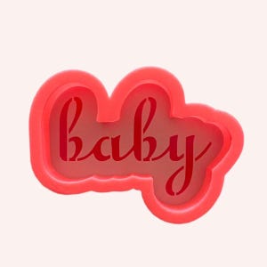 May include: A bright coral-coloured cookie cutter featuring a cursive script font that spells out the word baby. The object has a thick, rounded outer frame and a recessed inner section that forms the letters of the word in a connected style.