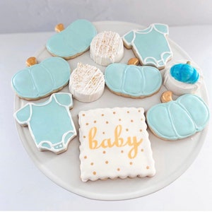 May include: A collection of decorated sugar cookies featuring light blue pumpkins with gold stems, light blue baby bodysuits with white piping, white round cookies with gold splatter, and a square white cookie with gold dots and the word baby.