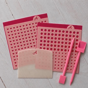 May include: Two rectangular pink stencils with heart and circle patterns, a translucent white scraper, and two pink tools. Each piece features a small tab with the letter A. The tools are marked with the text SASSY ART STENCILS in pink lettering.