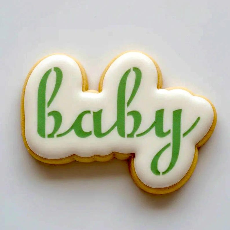 May include: A sugar biscuit shaped like the word baby in cursive script. The biscuit features a smooth white icing base with the word baby written in a vibrant green colour. The edges of the biscuit are defined by a thin golden brown crust.